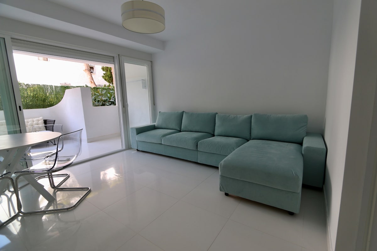 A bright living area features a spacious light blue sectional sofa and a round dining table with sleek transparent chairs. Large sliding glass doors provide access to a private outdoor space filled with greenery.
