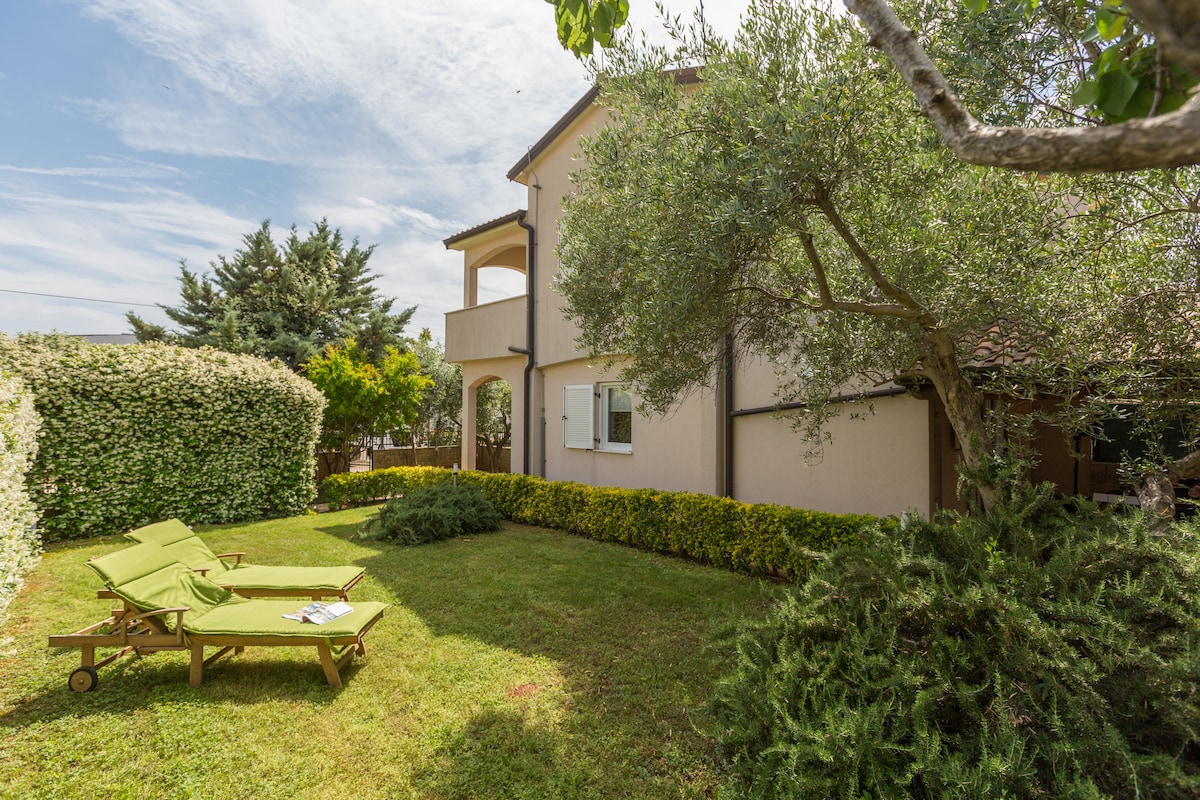 A well-maintained garden features two green lounge chairs positioned on a grassy area. Surrounding the space, various shrubs and trees create a natural boundary, enhancing privacy. The house's exterior is visible, showcasing a light-colored facade and small balconies on the upper floor.