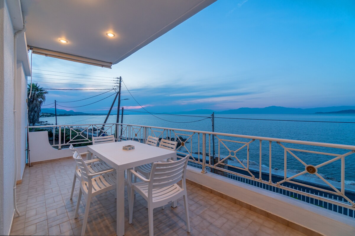 A spacious balcony with a dining table and white chairs provides an unobstructed view of the sea and distant mountains. The soft colors of the sky at dusk are visible, enhancing the serene coastal environment.
