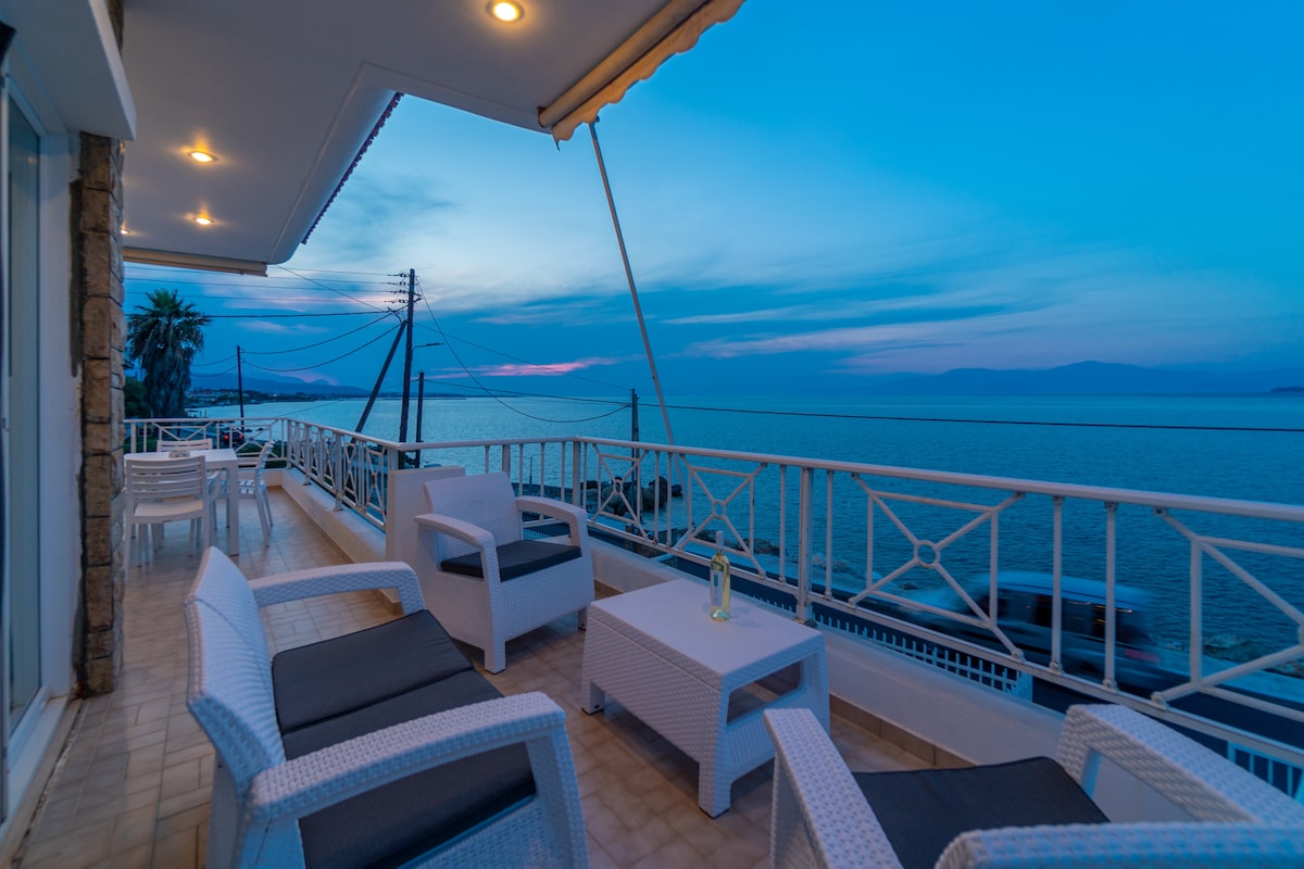 A spacious balcony is featured, equipped with stylish white seating and a small coffee table. The serene seascape is visible, showcasing a calming blue twilight sky. Power lines are seen in the distance, along with gentle waves lapping against the shore.