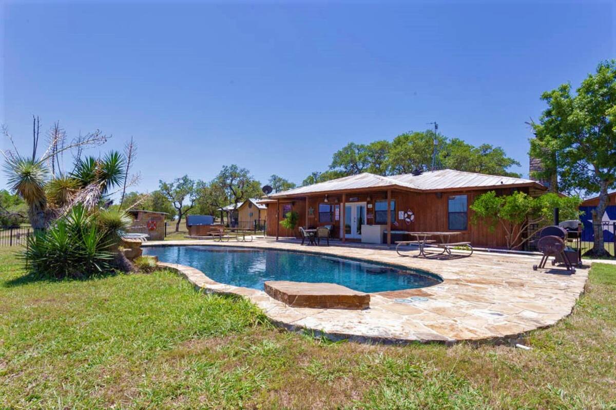 A spacious outdoor area features a clear blue pool surrounded by natural stone and lush greenery. A charming wooden home is positioned nearby, adorned with large windows and a covered porch. Comfortable lounge chairs are scattered around the pool, inviting relaxation.