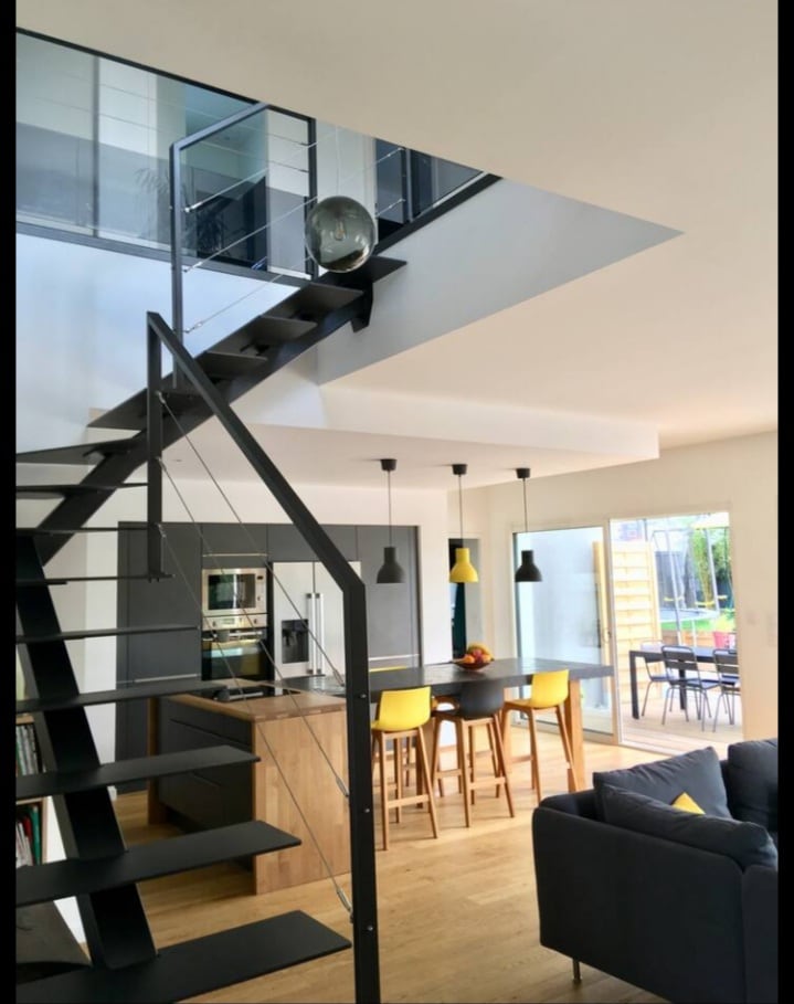 A modern interior showcases an open layout with a staircase featuring black railings leading to the upper level. An island kitchen bar with yellow chairs is visible, alongside sleek cabinetry. Large windows allow natural light to brighten the space, creating a welcoming atmosphere.