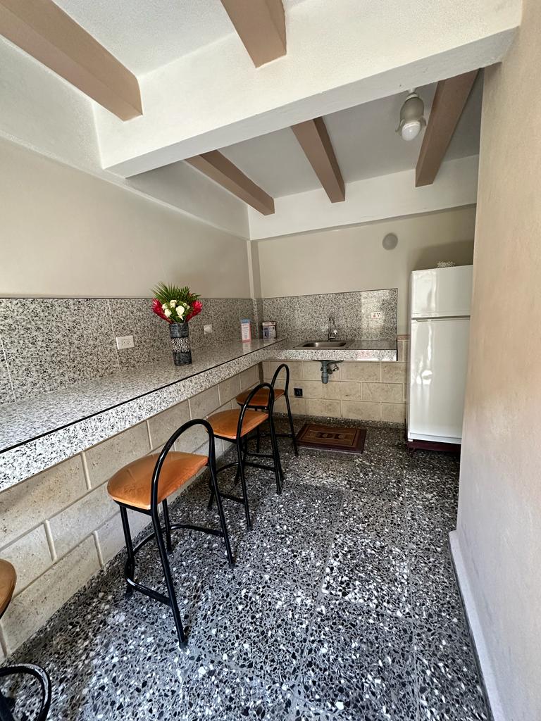 A common pantry area features a long countertop, equipped with various amenities. Three wooden stools are positioned along the countertop, while a white refrigerator stands against the wall. A flower arrangement adds a touch of color to the space, which is illuminated by natural light.