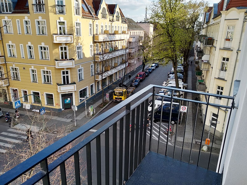 A view from the balcony overlooks a tree-lined street with parked cars. Yellow and cream-colored buildings are visible across the street, showcasing various balconies. Street signs and a crosswalk enhance the urban environment, while sunlight illuminates the scene.
