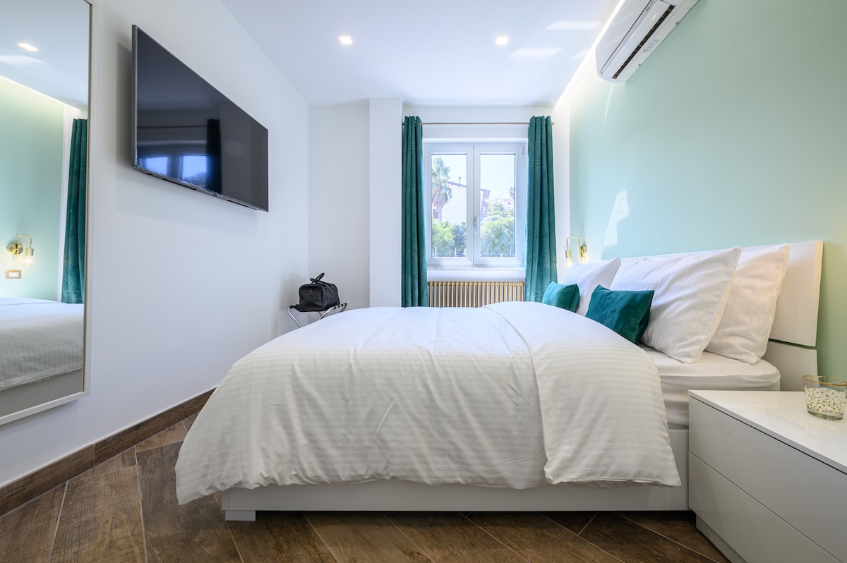 A well-lit bedroom features a spacious bed with crisp white linens and teal accent pillows. A large mirror reflects the room, while a mounted TV is positioned on the wall. The window is dressed with teal curtains, allowing natural light to enhance the space.