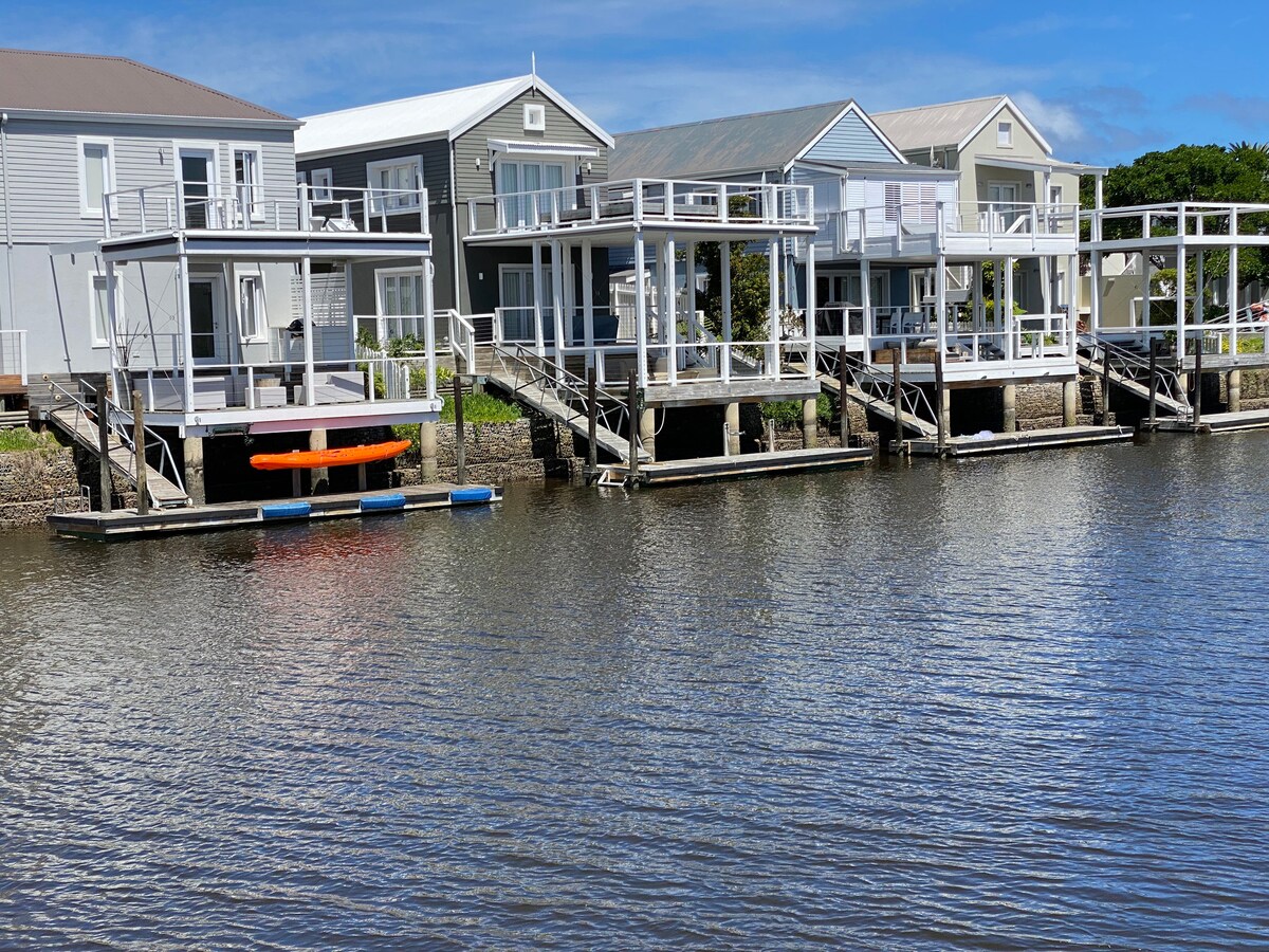 Multiple waterfront houses with decks are positioned along a calm river. Bright orange kayaks and a blue floating dock are visible nearby. The setting is enhanced by clear skies and interspersed greenery.