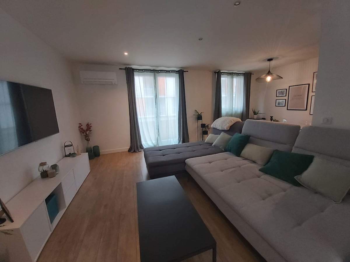 A modern living area is depicted with a sectional sofa featuring soft cushions, positioned adjacent to a sleek black coffee table. Large windows are dressed with light curtains allowing natural light to filter in. A flat-screen TV is mounted on the wall, enhancing the contemporary design.