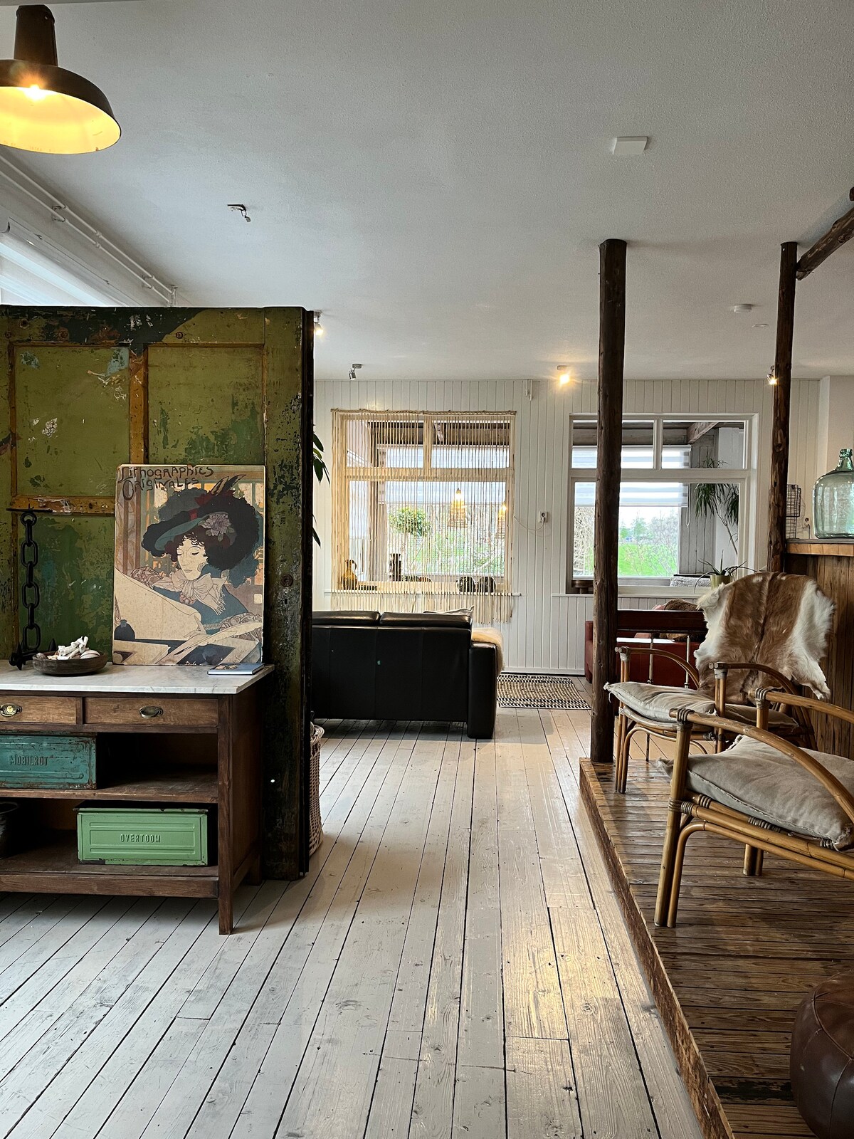 A living space showcases vintage-inspired furniture, including a wooden desk and a stylish chair. Large windows provide natural light, illuminating the cozy seating area at the far end. Unique decor elements, such as an artistic print and a rustic wall, enhance the design of this inviting apartment.