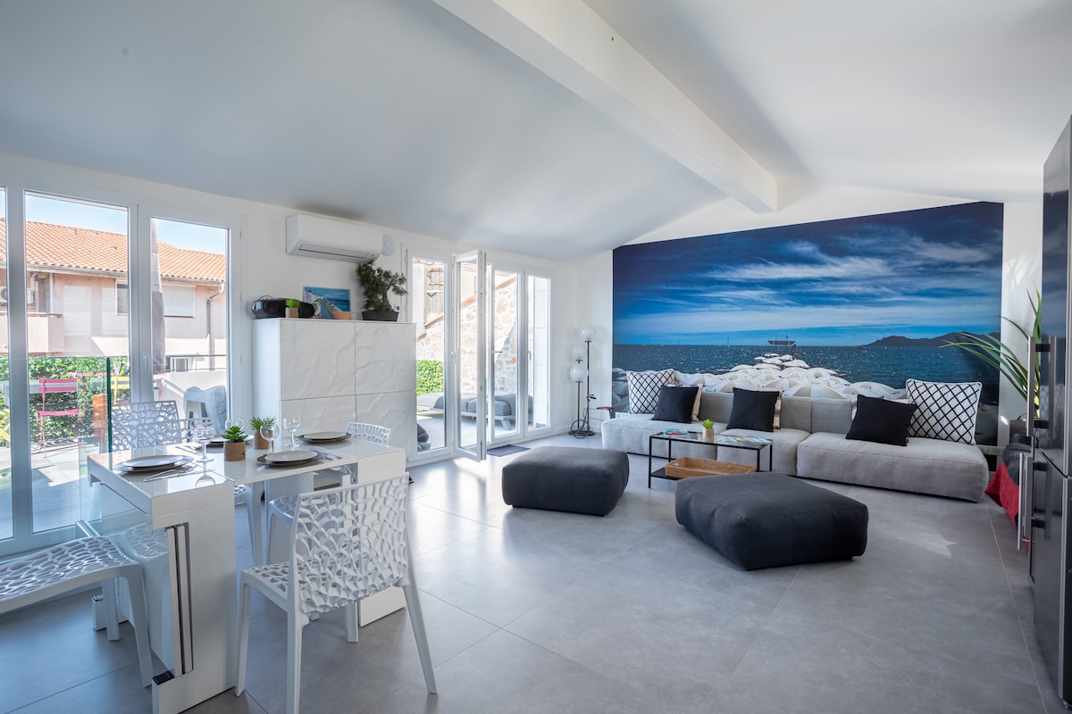 The spacious living area is filled with natural light from large windows. A modern, comfortable sofa is complemented by soft, low seating. A dining table set for meals stands adjacent, while a wall mural depicting a serene coastal scene adds visual interest.