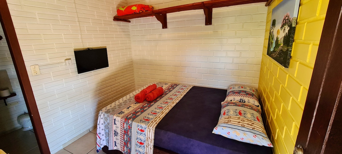 A cozy couple's suite features a spacious bed adorned with colorful pillows and rolled towels. A small television is mounted on the wall, and bright yellow walls add a cheerful tone. Natural light illuminates the room, highlighting the clean, tiled flooring.