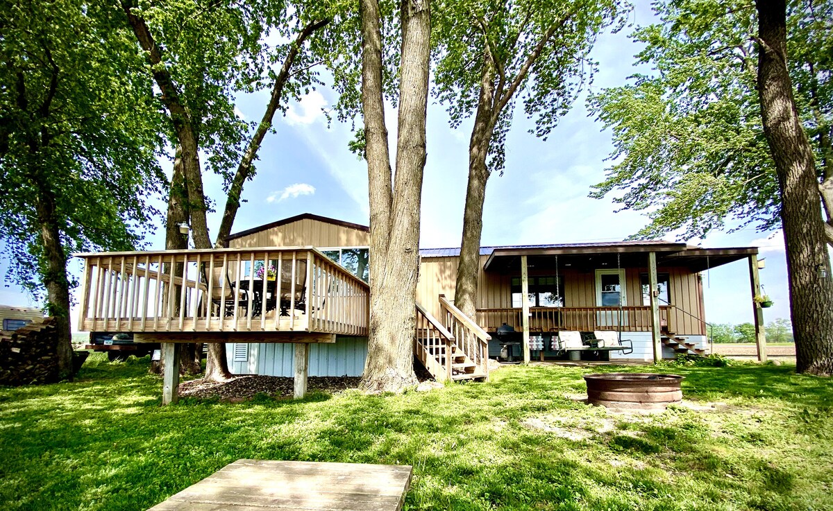 A spacious cabin is nestled among tall trees, showcasing a large deck with seating and a campfire ring below. Green grass surrounds the structure, inviting relaxation in a serene natural setting. The exterior features a combination of wood and light-colored siding.