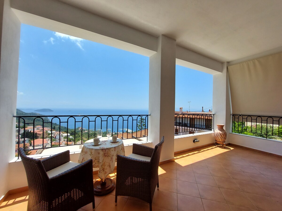 An outdoor balcony area is featured, offering expansive views of the Aegean Sea and surrounding hills. Two comfortable chairs are positioned around a small table, complemented by a terracotta pot. Bright blue skies and a distant coastline enhance the serene atmosphere.