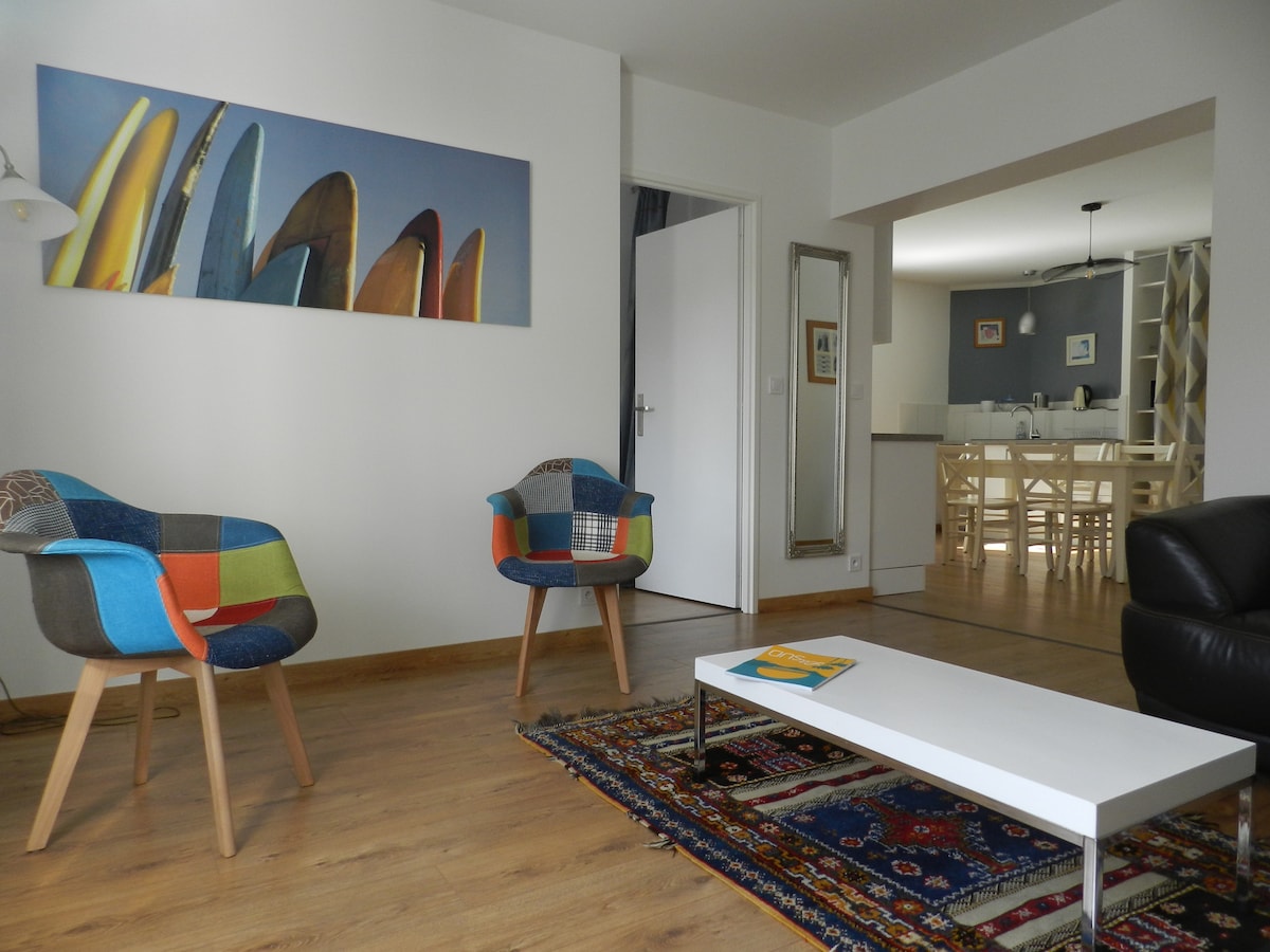 A bright living area features two colorful armchairs and a low coffee table atop a patterned rug. A large canvas depicting surfboards is displayed on the wall, while an open layout allows visibility of the dining area and kitchen in the background.