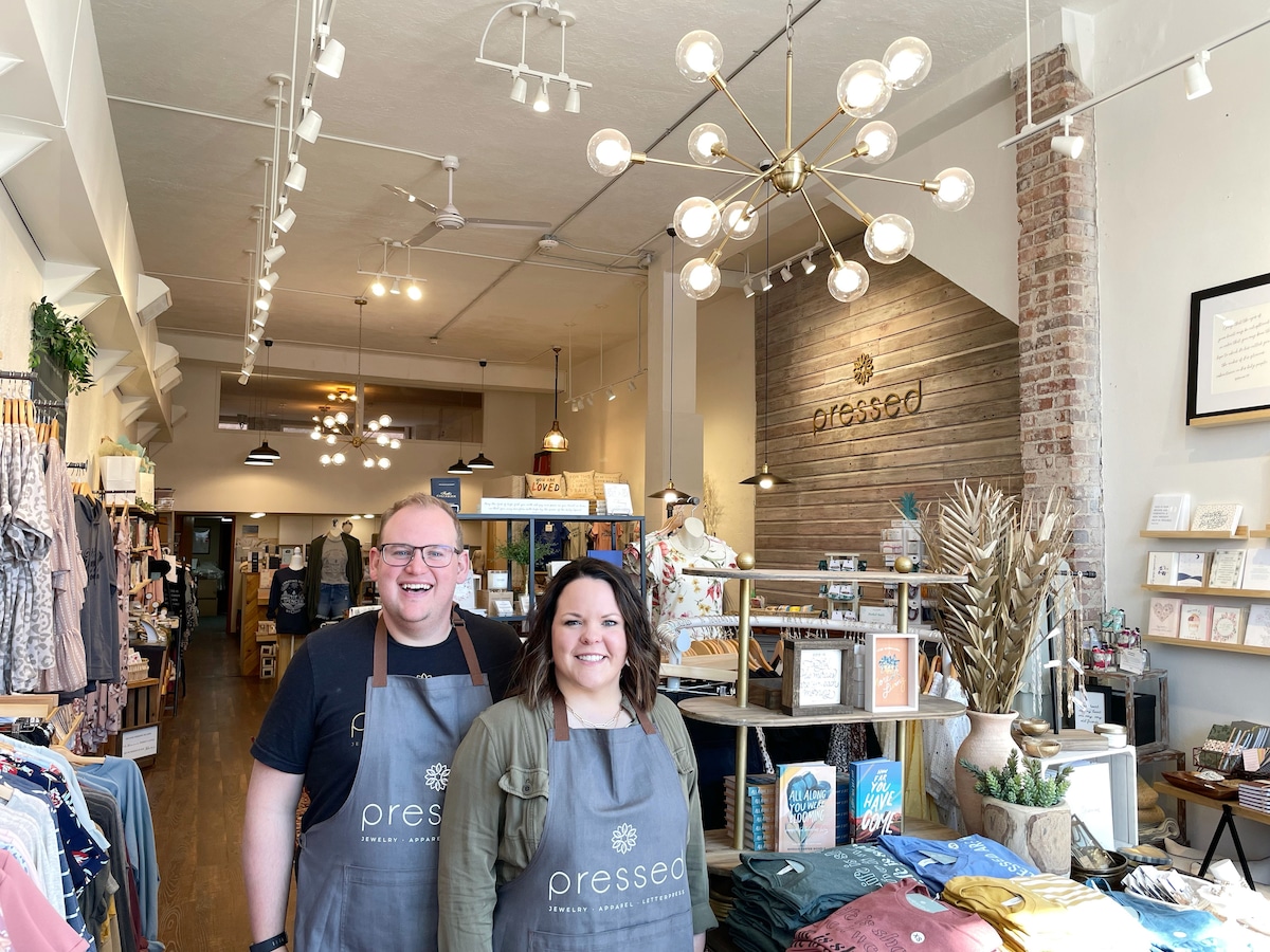 The interior of the retail store, Pressed, is showcased, featuring an open layout with displays of clothing and merchandise. Hanging light fixtures illuminate the space, while wooden and brick accents add character. Two individuals stand in the foreground, wearing branded aprons.