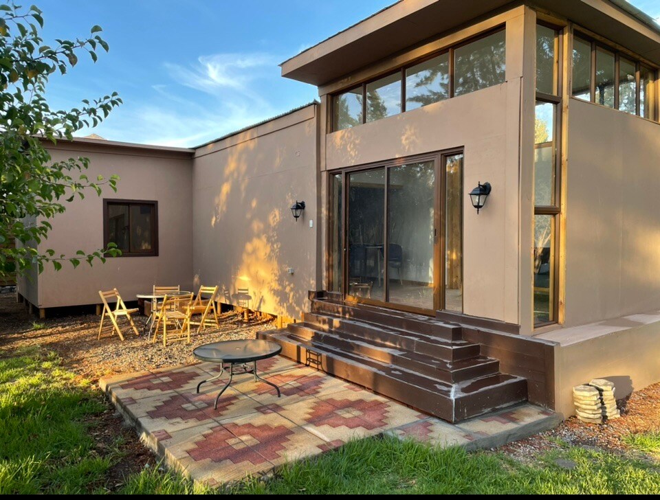 A modern cabin features large windows allowing natural light to illuminate the interior. Steps lead up to the entrance, with a patio area furnished with a round table and several folding chairs. Lush greenery surrounds the outdoor space, contributing to a calm environment.