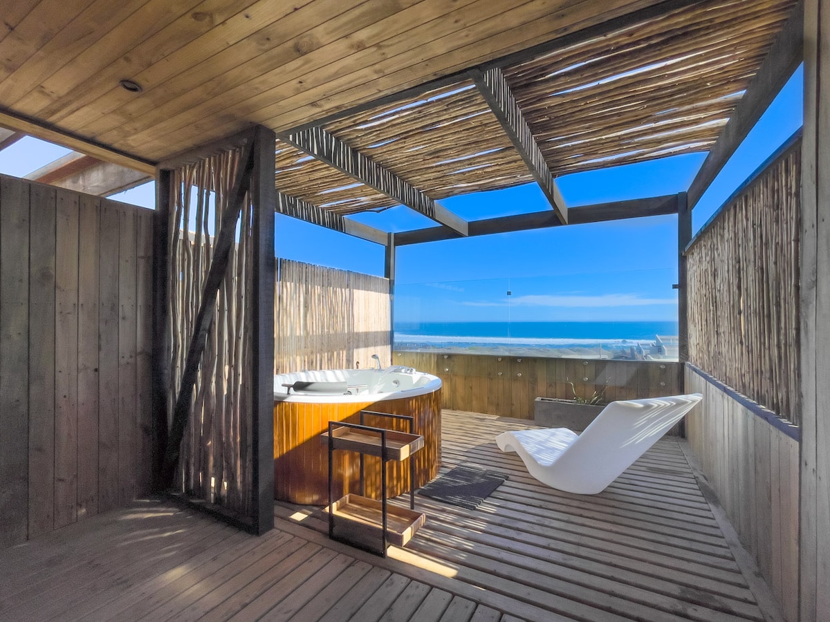 A private hot tub is surrounded by wooden decking, offering an unobstructed view of the ocean. A single lounge chair is positioned nearby under a shaded pergola, allowing for relaxation amidst open-air surroundings. Natural light highlights the simple, rustic design of the space.