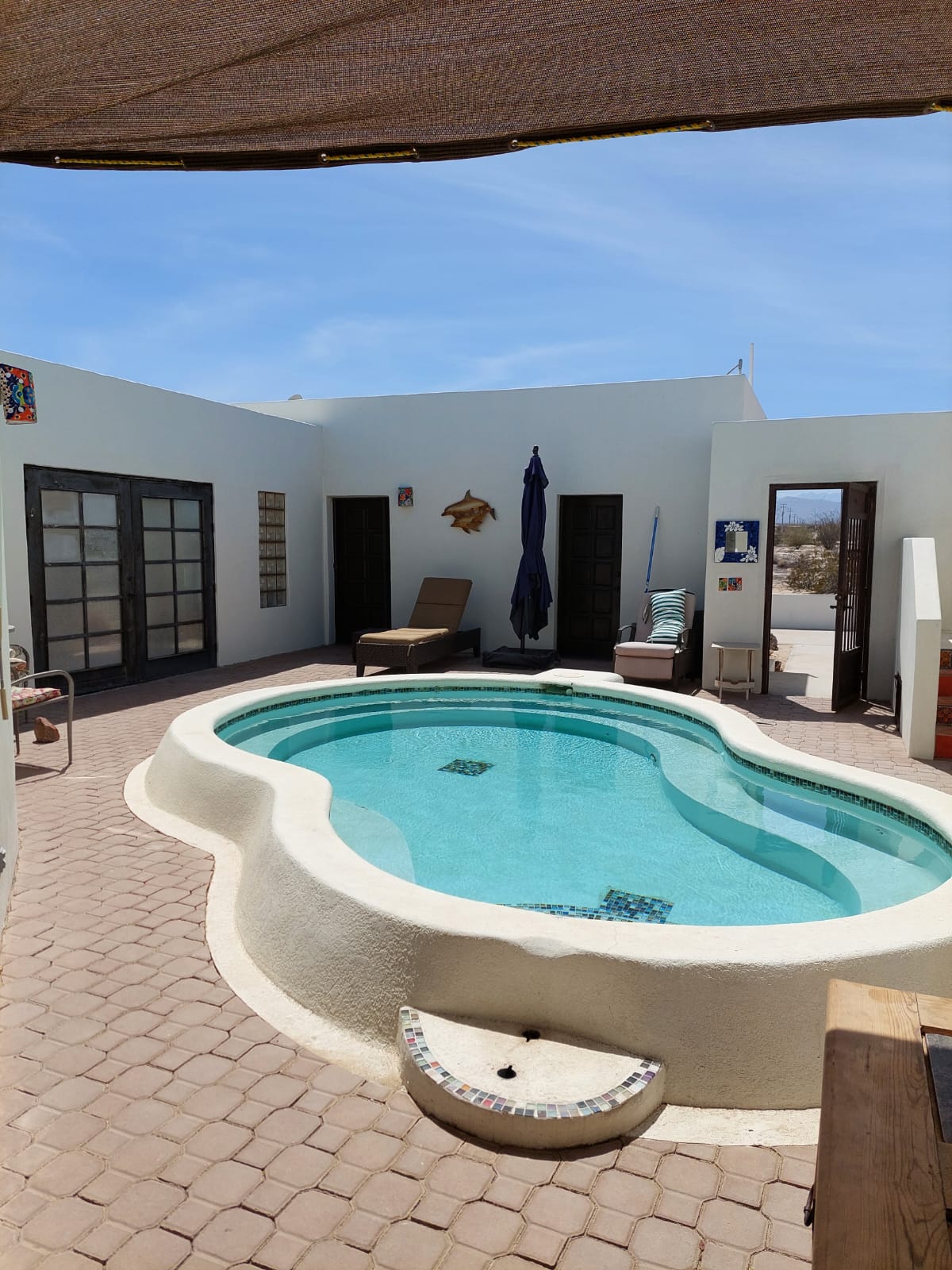 An inviting private pool is surrounded by stone-paved flooring, with a comfortable lounge chair positioned nearby. Light blue water reflects the sunlight, while tall, dark panels serve as entrances to adjacent spaces. The surrounding walls are painted in a warm, light color.