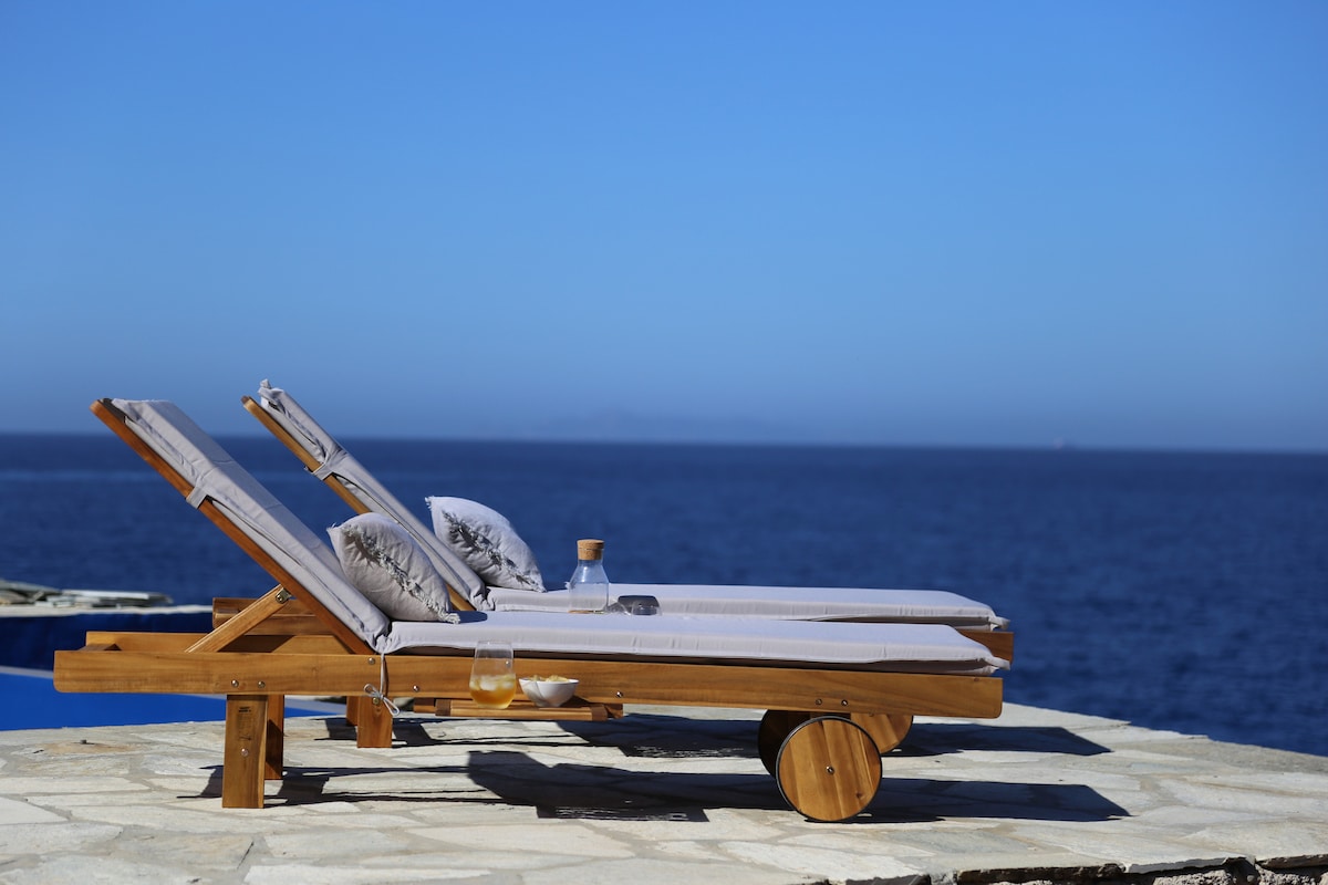 Two wooden sun loungers are positioned on a stone terrace overlooking the expansive blue sea. Each lounger is equipped with soft gray cushions and a small glass bottle is placed between them. The clear blue sky complements the serene ocean view.