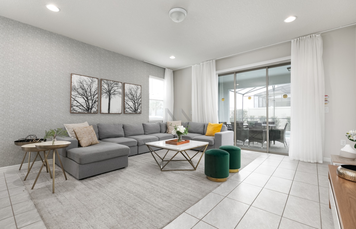 A spacious living area features a light grey sectional sofa complemented by two green stools and a pair of decorative tables. Large windows provide ample natural light, with sheer curtains softly framing the view. Elegant wall art depicting trees adds a touch of style to the room.