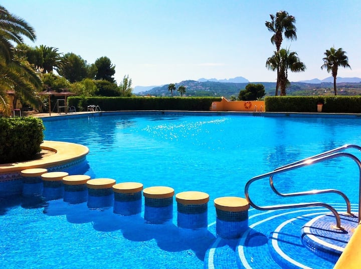 A large swimming pool is surrounded by palm trees and greenery, with clear blue water reflecting sunlight. Circular seating areas are positioned along the pool’s edge, leading towards a gentle staircase. The distant mountains are visible in the background against a bright blue sky.