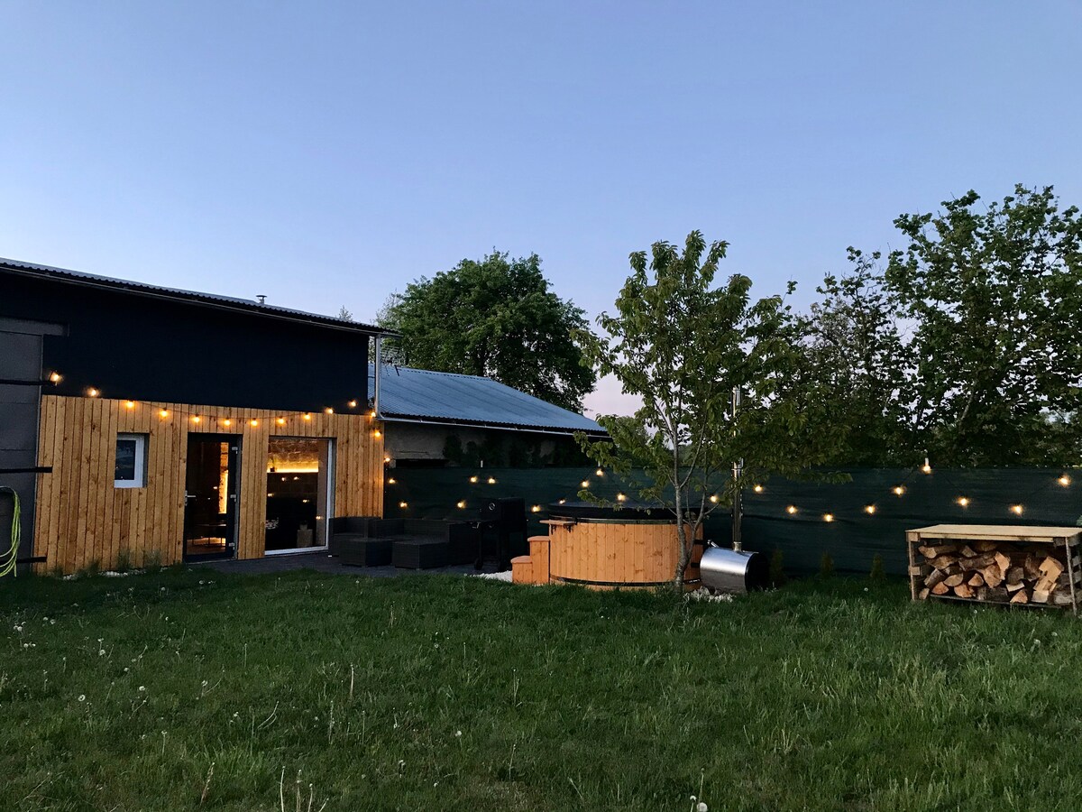 An outdoor area features a well-maintained green lawn illuminated by string lights. In the foreground, a wooden hot tub is positioned beside a cozy seating arrangement. A stack of firewood is visible next to a small fire pit, surrounded by lush trees.