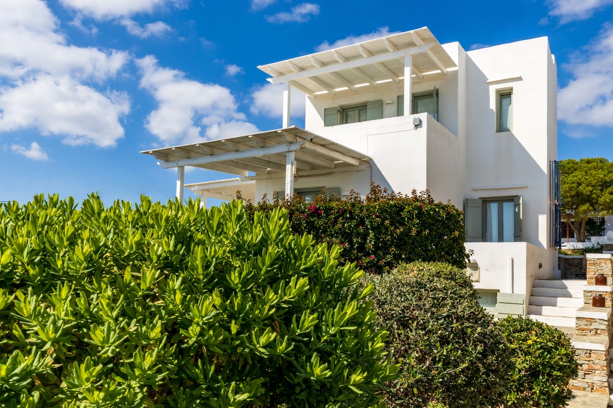 A modern two-story house featuring a clean white facade is surrounded by lush greenery. A spacious terrace is visible on the upper level, offering views of the sky. Large windows are present, allowing natural light to enter the interior spaces.