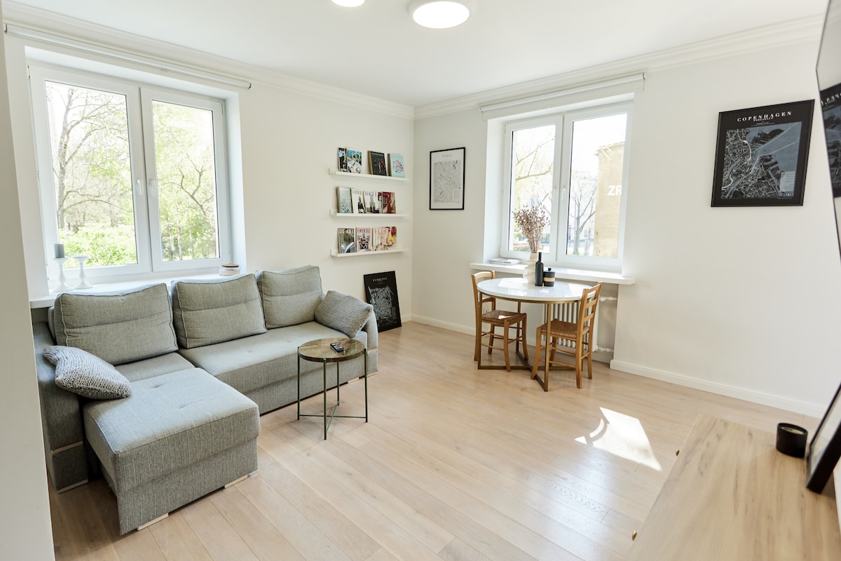 The living room features a comfortable gray sofa with a chaise lounge, complemented by a small glass side table. A round dining table is placed nearby, accompanied by two chairs. Natural light enters through two large windows, illuminating the warm wooden floor.