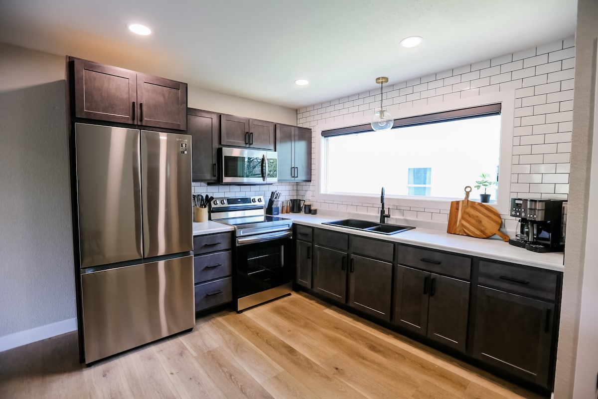 A modern kitchen equipped with stainless steel appliances, including a refrigerator and oven. The sleek cabinetry offers ample storage space, while a large window provides natural light. A minimalist sink is complemented by essential kitchen tools and an attractive cutting board.