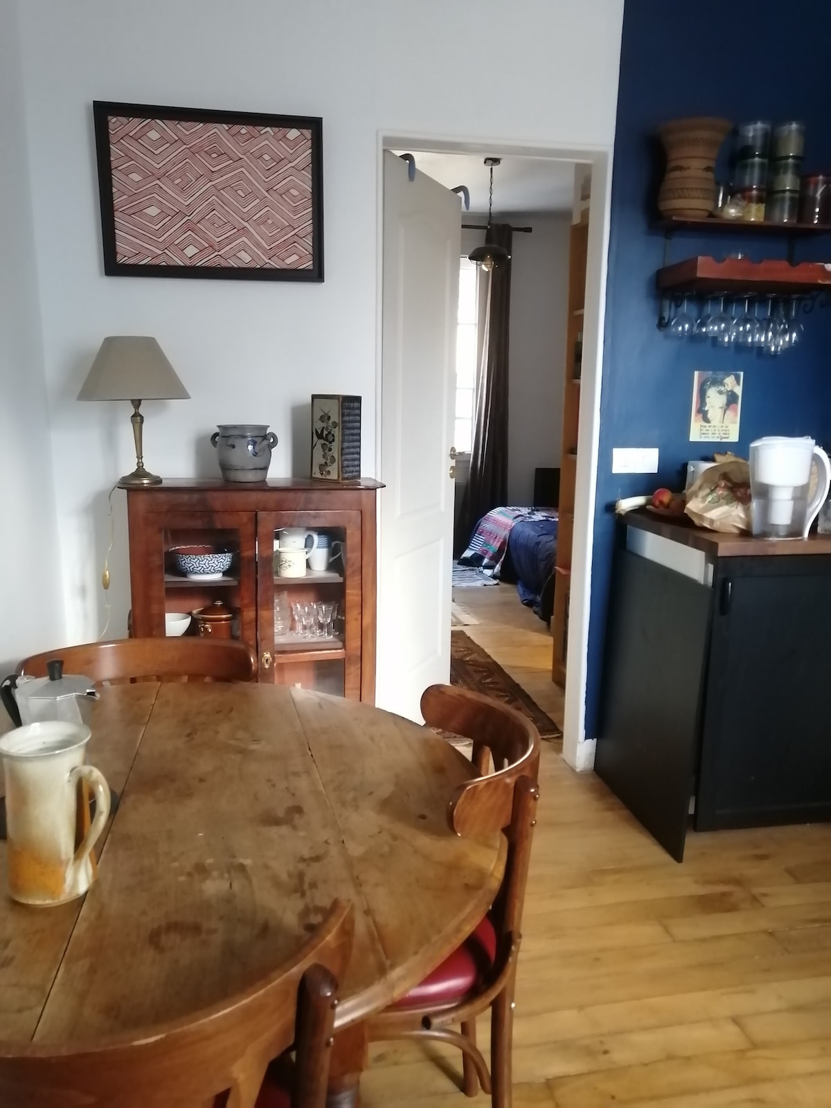 A dining area features a round wooden table surrounded by four chairs. A lamp stands on a side table, complemented by a wall-mounted shelf displaying glassware. An open door reveals glimpses of a bedroom with blue walls, while a black cabinet and kitchen items are visible nearby.