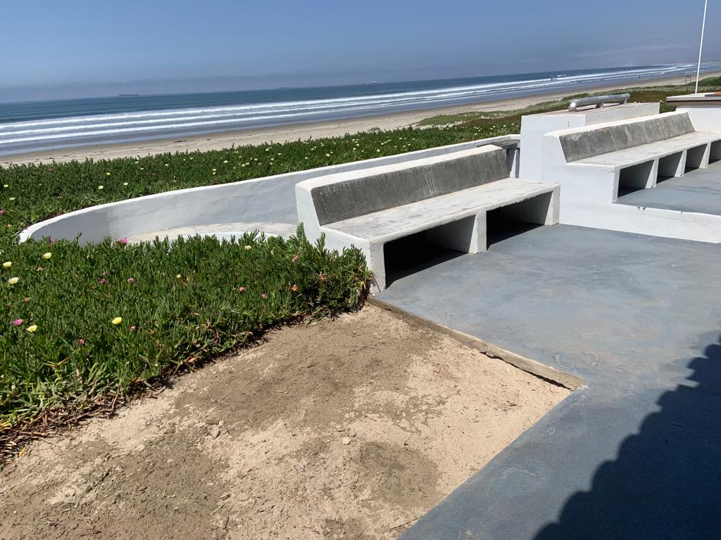 A paved outdoor area features built-in seating along a curved wall, overlooking a sandy beach. Green landscaping with flowering plants borders the space, while the waves gently lap against the shore in the distance under a clear sky.