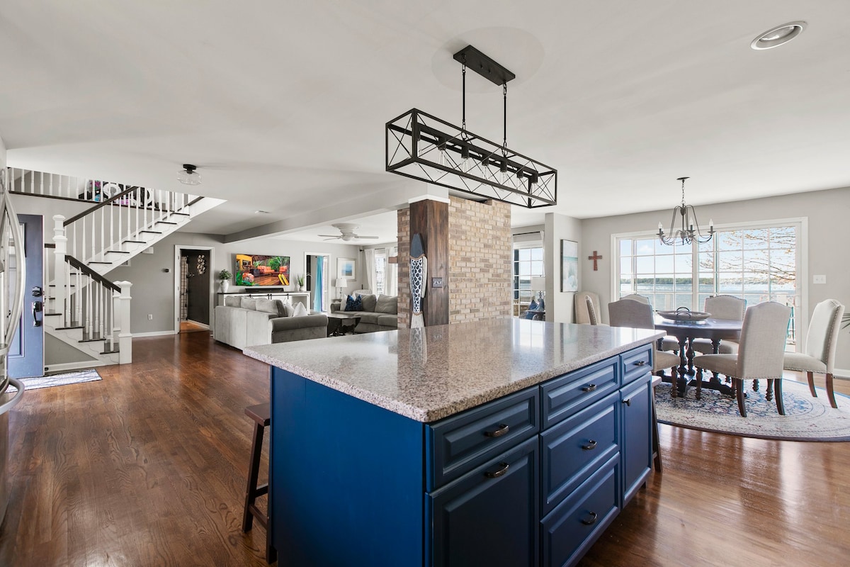An open floor plan is revealed, featuring a spacious kitchen island with blue cabinetry and a granite countertop. A large dining area is visible in the background, complemented by natural light from nearby windows, while a staircase leads to the upper level.