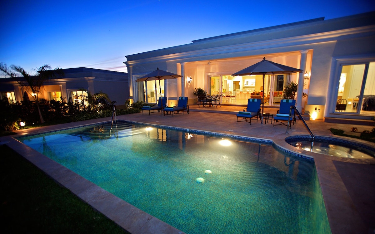 An inviting outdoor space features a glistening pool illuminated under the evening sky. Two shaded lounge areas with blue chairs are positioned by the pool, offering a relaxing atmosphere. Large glass doors reflect the warm light from the interior, enhancing the serene ambiance.