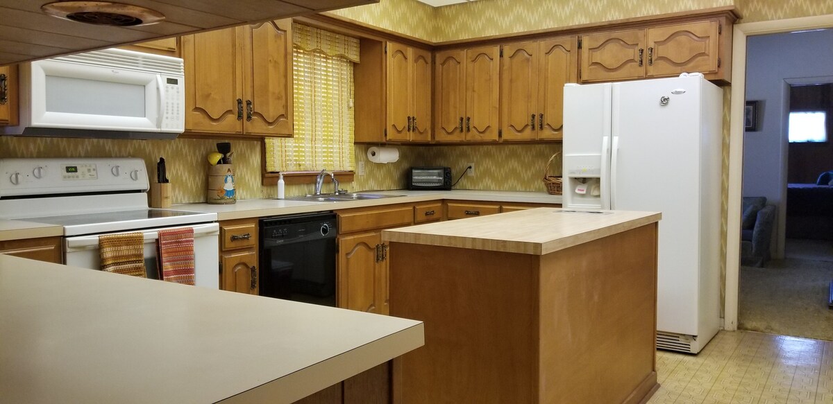 The kitchen features wooden cabinetry and a spacious countertop. Modern appliances include a microwave, oven, and refrigerator. A window with blinds allows natural light and offers a view of the outdoor area. The layout provides an open feel connecting to the dining space.