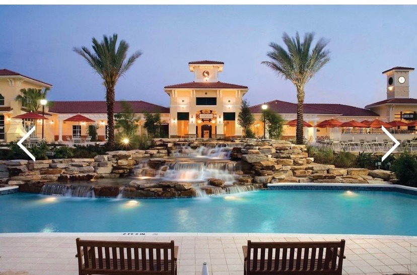 A resort-style pool is highlighted by a cascading waterfall and surrounded by palm trees. Comfortable seating is positioned along the edge of the pool. The inviting atmosphere is complemented by the brightly lit resort building visible in the background.