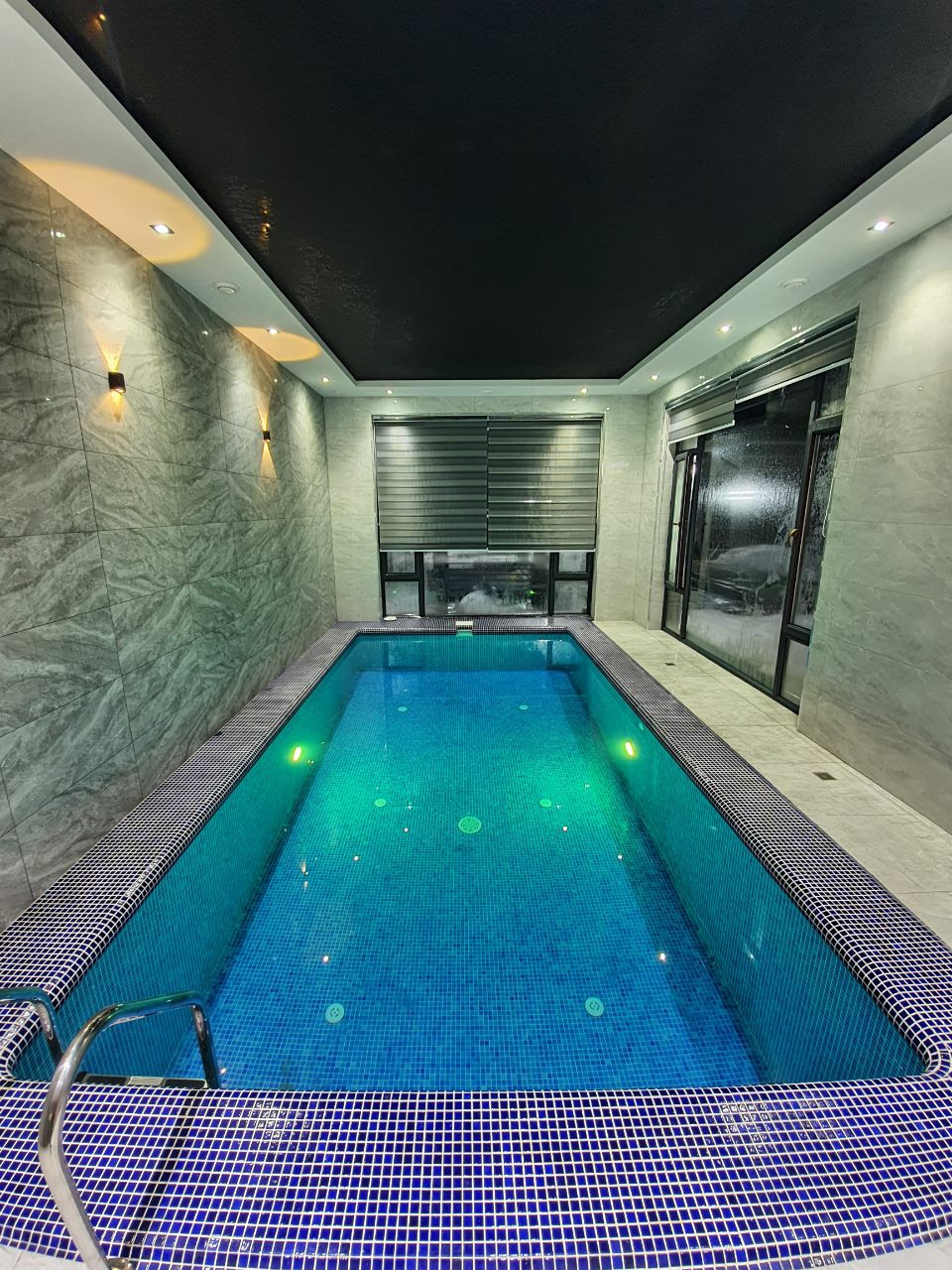 An indoor swimming pool is featured, with sparkling blue tiles and modern lighting accents. Large windows allow natural light to filter in, creating a serene atmosphere. The pool includes steps for easy access and is surrounded by sleek stone walls.
