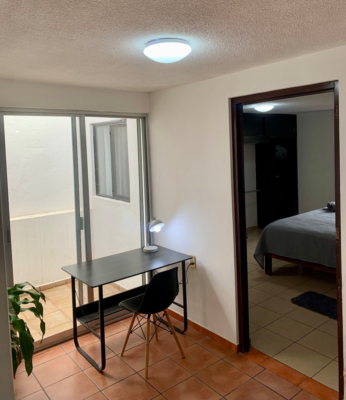 A well-lit corner features a simple desk with a black chair and a small lamp, positioned near glass doors that lead to a balcony. The doorway opens to a spacious room with a neatly made bed visible in the background.