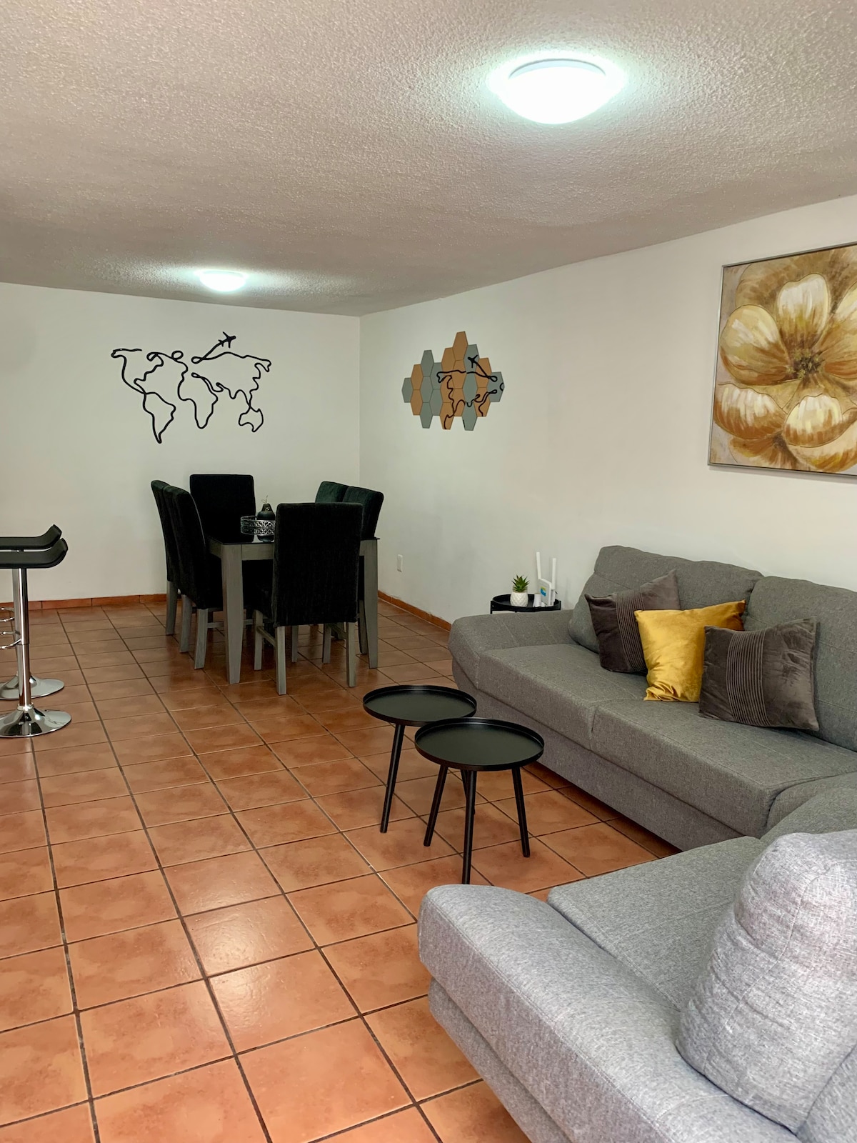 A spacious living and dining area is shown, featuring a gray sofa with decorative cushions and two small black side tables. A wooden dining table is surrounded by six chairs, with wall art displaying a world map and a floral piece enhancing the decor.