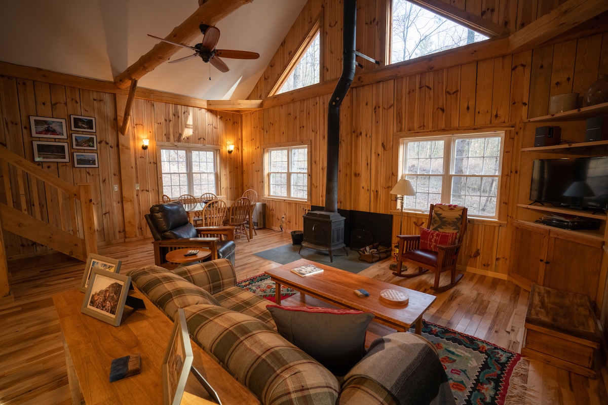 A warm living area features wooden walls and beams, with large windows allowing natural light. A cozy sofa is arranged around a wooden coffee table, while a small table with wicker chairs sits in one corner. A ceiling fan enhances the airy atmosphere.