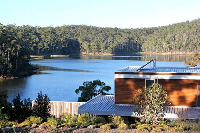A modern home is situated near a serene lake, surrounded by lush green trees. The water reflects the tranquil landscape, enhancing the peaceful setting. The structure features a contemporary design with wooden accents, and a landscaped area is visible in the foreground.