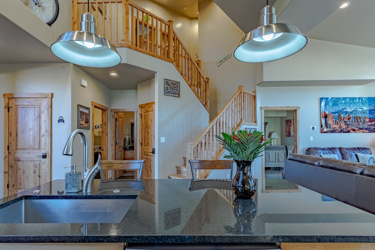 A view showcasing a modern kitchen island with a dark granite countertop and a stylish faucet. Bright pendant lights hang above, illuminating the space. The open concept design leads to a cozy living area and wooden staircase, enhancing the airy feel of the home.