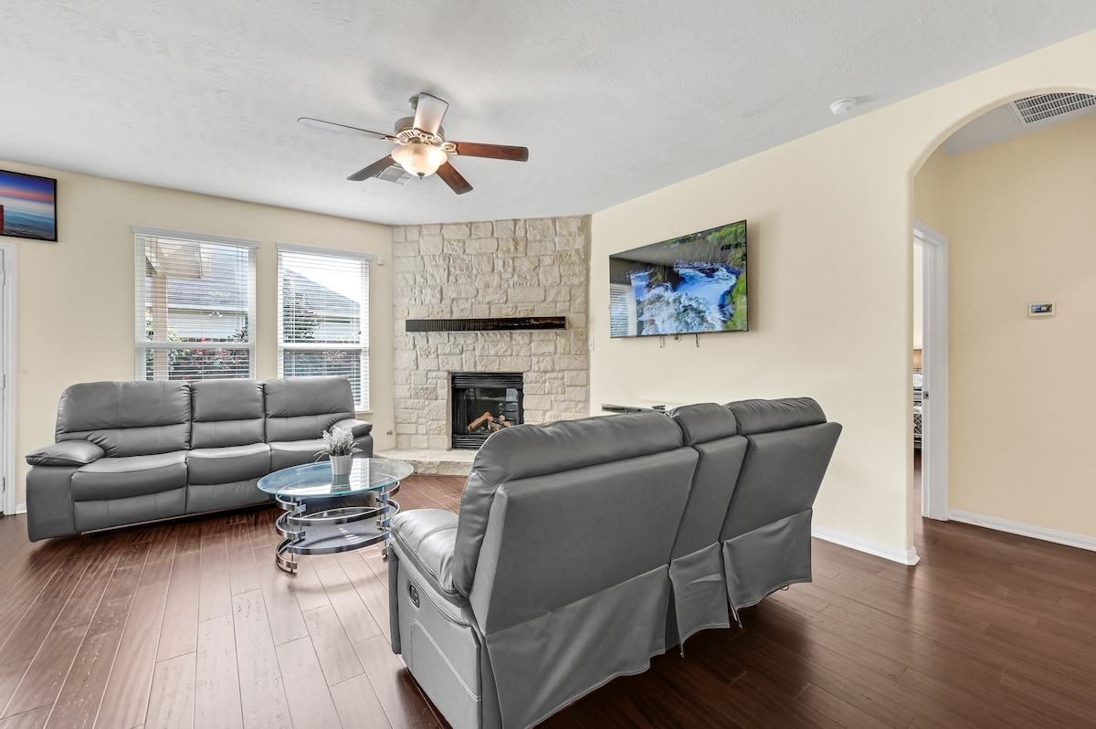 The living room features a gas log fireplace set within a full stone wall. Comfortable gray sofas surround a glass coffee table, while large windows allow natural light to fill the space. A mounted television is positioned above the fireplace, enhancing the room’s functionality.