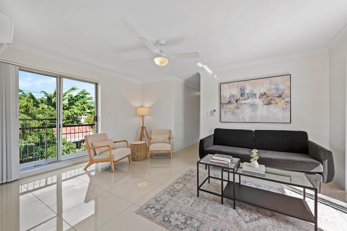 The living room is bright and airy, featuring large windows that allow natural light to fill the space. A comfortable black sofa is positioned in front of a modern coffee table, accented by a decorative plant. Two light armchairs and a lamp provide additional seating and warmth.