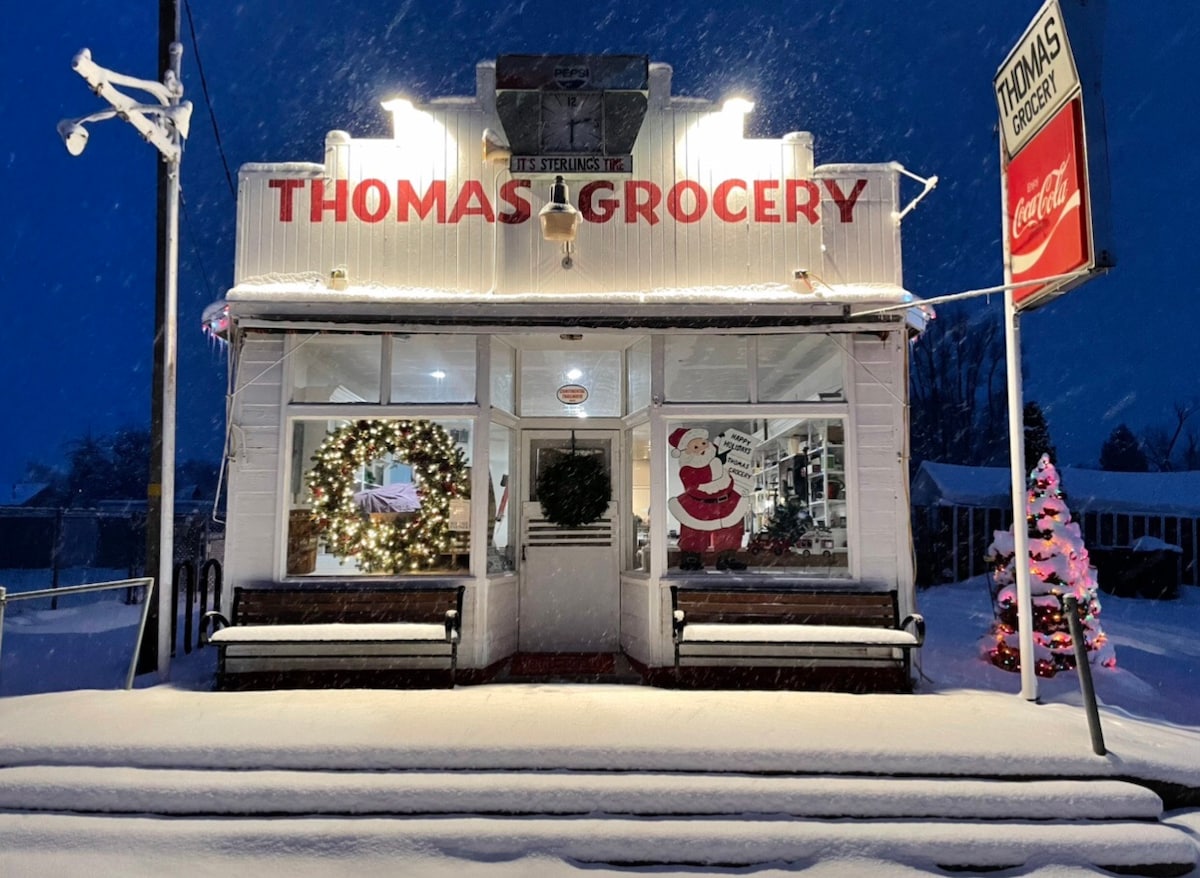 The exterior of Thomas Grocery is shown against a snowy backdrop, featuring a wreath and festive decorations. Two wooden benches sit at the front under a covered awning. The store's sign and illuminated tree add seasonal charm to the serene winter scene.