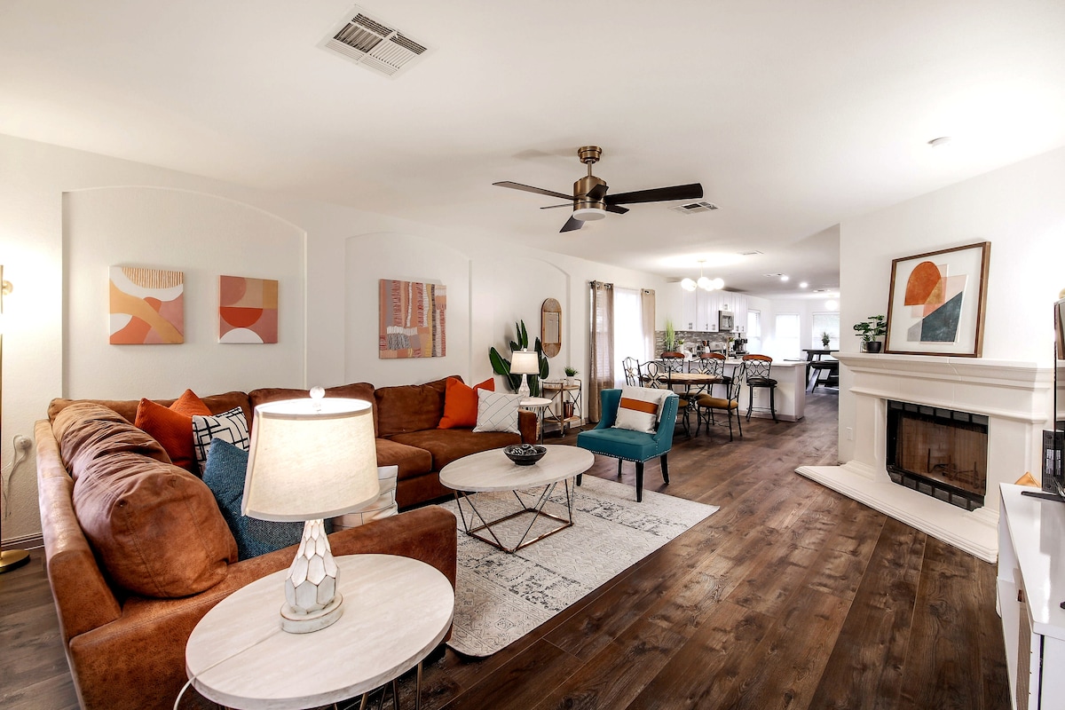 A spacious living room is furnished with a modern brown sectional sofa and a light-colored coffee table. Accents of blue and orange pillows add color to the area. A ceiling fan and wall art complement the room, which connects to a dining space visible in the background.