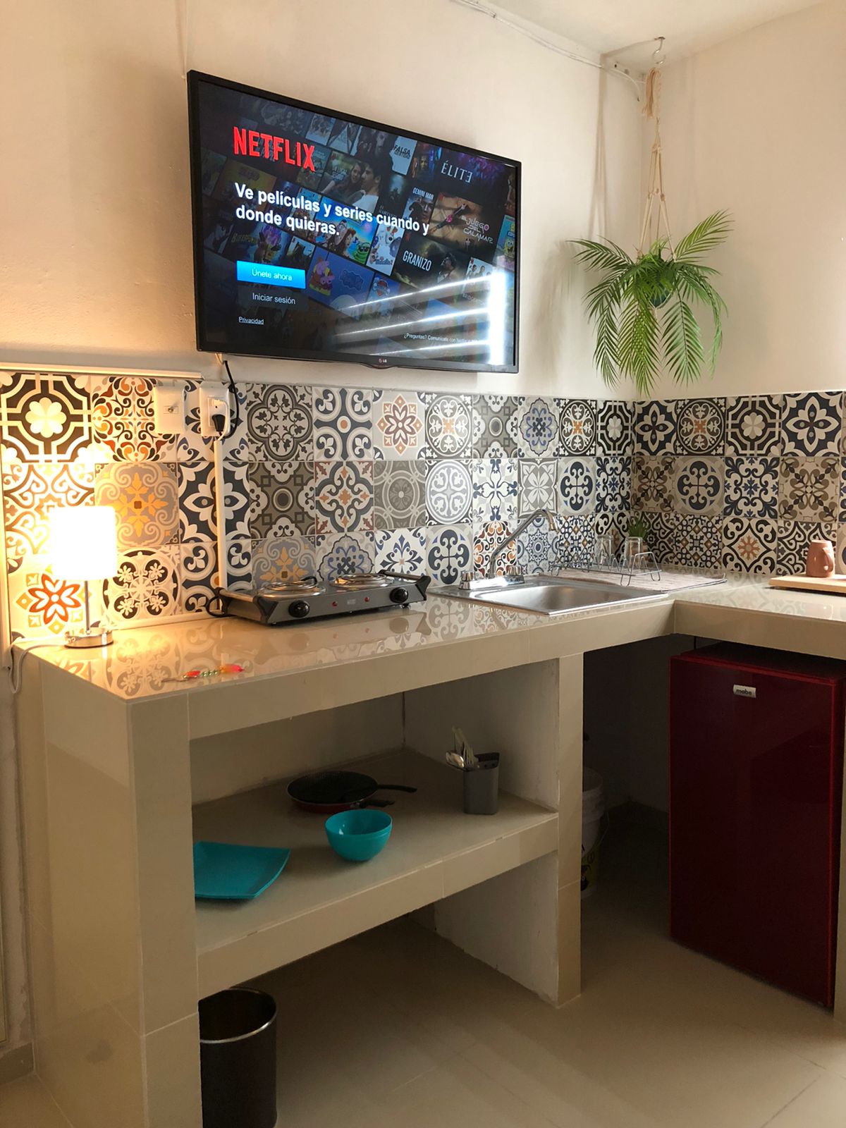 The kitchen features a modern layout with decorative tile backsplash and an overhead flat-screen television. A small cooking area with a stove and sink is present, alongside a compact refrigerator. A plant adds a touch of greenery to the space, and various kitchen items are arranged on the countertop.