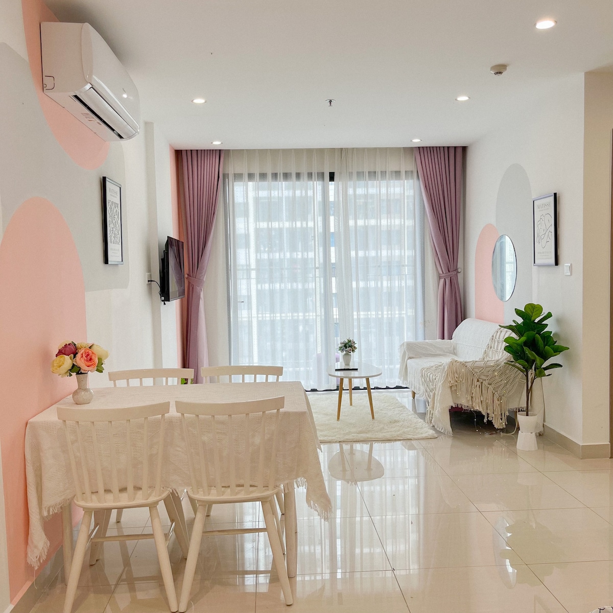 A bright living area features a white dining table set with four chairs, complemented by a cozy couch draped in a soft blanket. Floor-to-ceiling windows are adorned with sheer curtains, allowing natural light to fill the space. A small table is positioned nearby with a decorative plant.