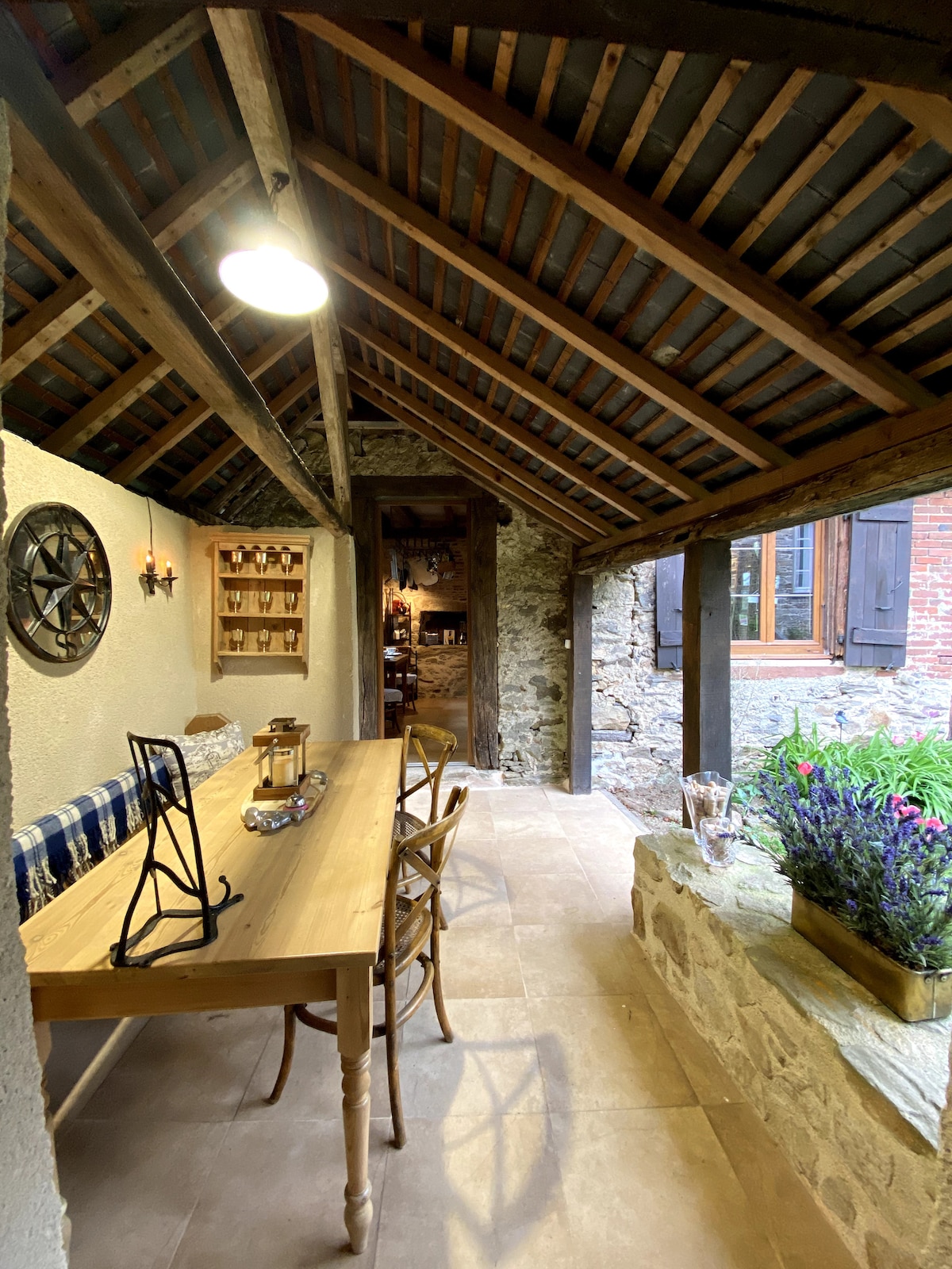A rustic dining area features a long wooden table surrounded by chairs, illuminated by warm overhead lighting. Exposed wooden beams and stone walls enhance the natural charm, while greenery peeks through a window leading to the outdoor patio.