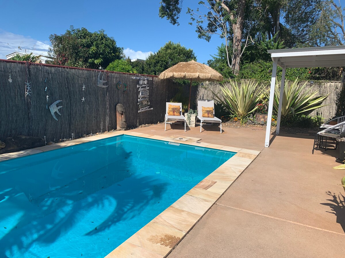 A serene pool area features a turquoise magnesium pool surrounded by a spacious patio. Two lounge chairs are positioned under a thatched umbrella, offering a shaded spot for relaxation. Lush greenery and tall plants create a private, tranquil setting.
