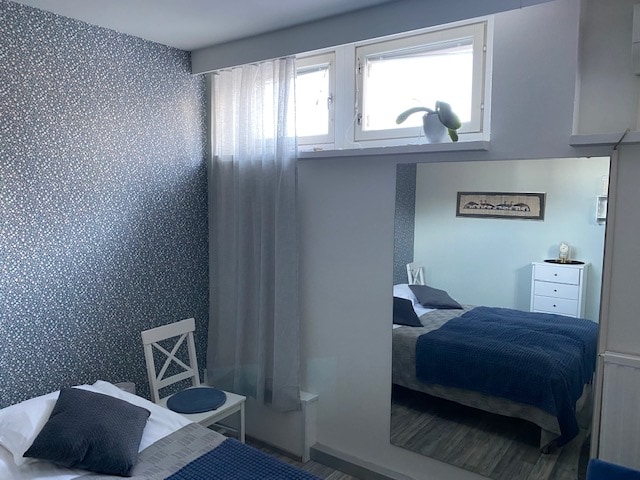 A bright bedroom features a window draped with a sheer curtain, allowing natural light to fill the space. A spacious bed is adorned with blue bedding and decorative pillows. A mirror reflects the room, which includes a white dresser and a small chair in the corner.