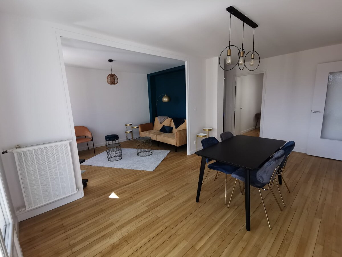 A spacious living area features a sleek wooden floor and a light color scheme. A dark table surrounded by blue chairs sits in the foreground. A cozy seating arrangement with a yellow sofa and modern lighting is visible in a separate space, framed by the bright walls.